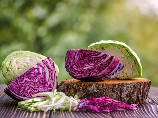 Cavolo cappuccio bianco e rosso, tagliato a spicchi e sminuzzato, su un tavolo di legno con sfondo verde sfocato.